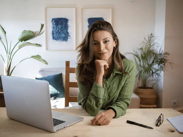 Een persoon zit aan een houten bureau met een laptop en een notitieboekje, met kamerplanten op de achtergrond.
