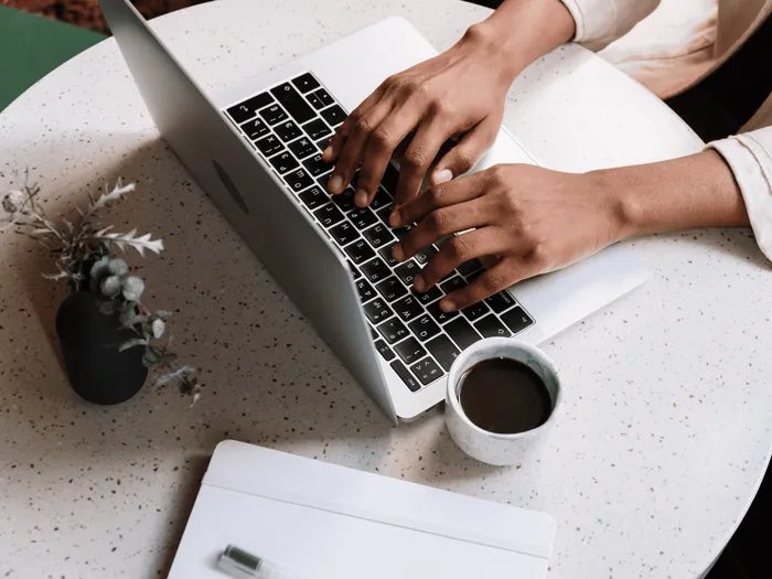 RS-blog-payroll-update-augustus-2026 Iemand die aan een tafel zit te werken, typend op een open laptop met de handen boven het toetsenbord, met een mok koffie en een klein potplantje ernaast.