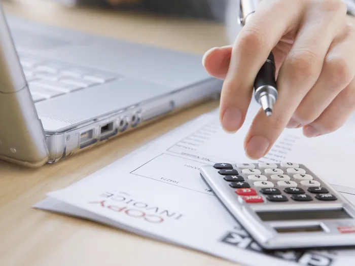 RS-blog-Facturatie Een hand die een pen vasthoudt boven een rekenmachine die op een factuur ligt, met op de achtergrond een laptop.
