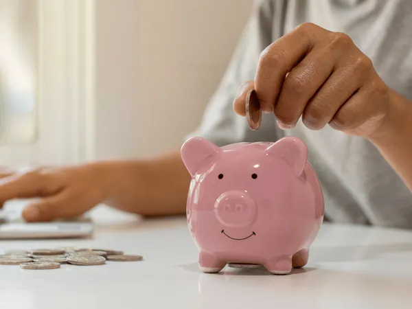 Een hand laat muntjes in een klein roze spaarvarkentje op een tafel vallen.