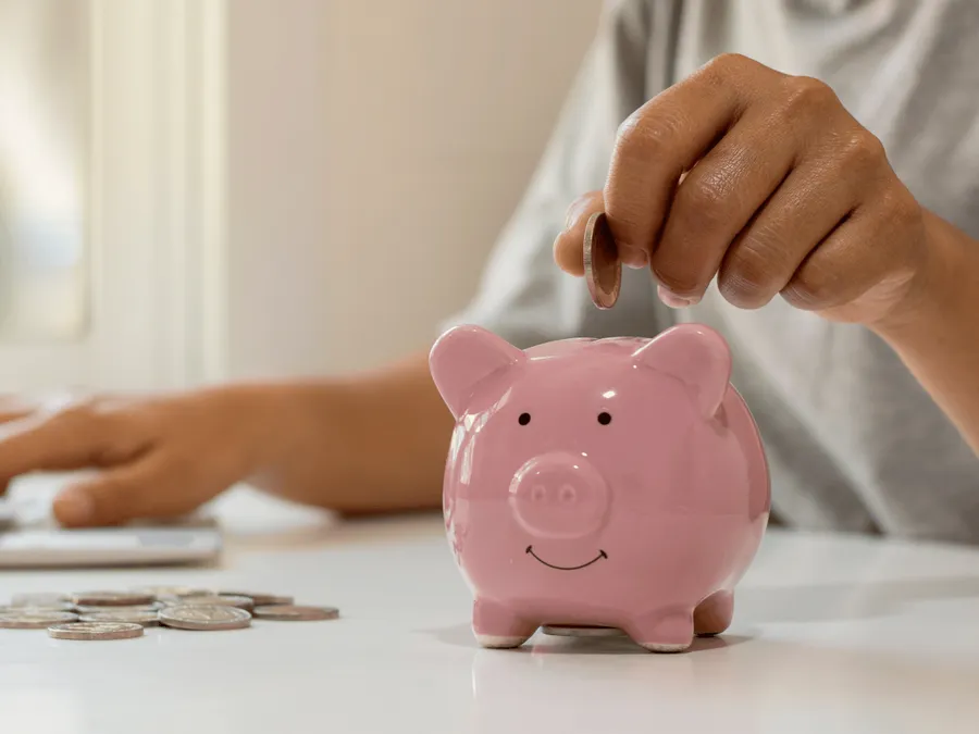 Een hand laat muntjes in een klein roze spaarvarkentje op een tafel vallen.