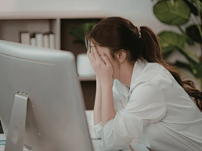 RS-blog-personeelsmutaties Een persoon met een bril die zijn gezicht met zijn handen bedekt, zittend voor een computerscherm in een kantooromgeving.