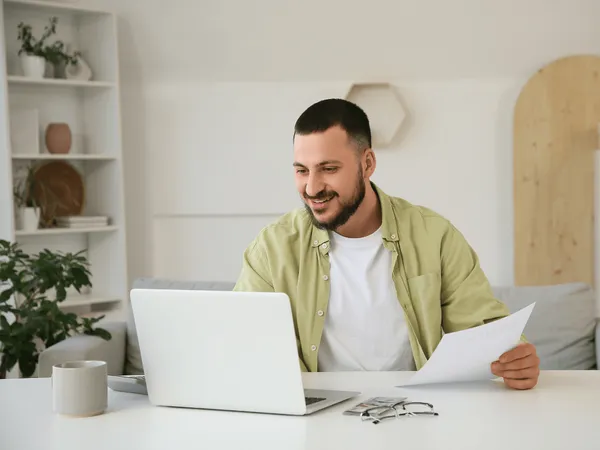 Een persoon in een lichtgroen shirt zit aan een bureau met een laptop en papieren, kijkt naar het scherm en glimlacht.