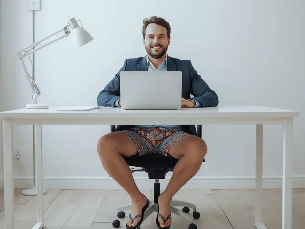 Een man in een colbert en een kleurrijke korte broek zit in slippers achter een bureau met een laptop.