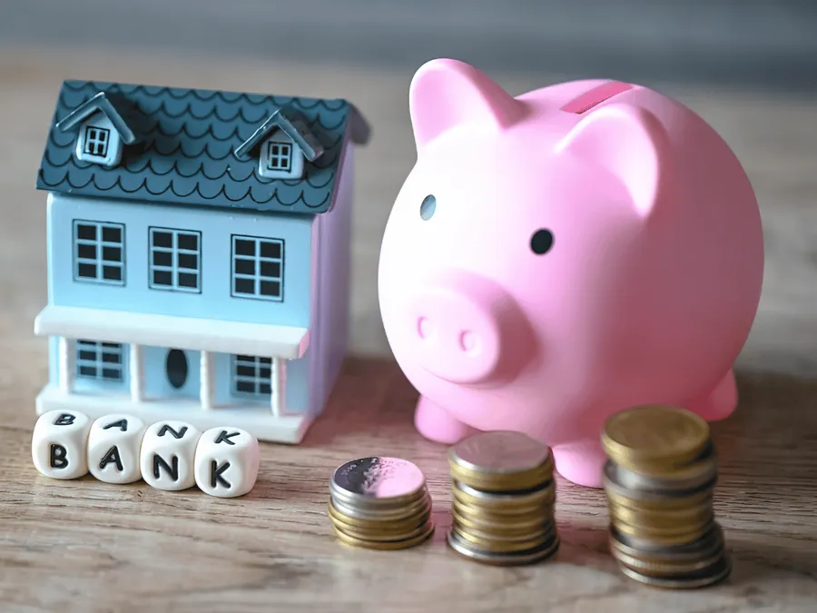 Een roze spaarvarken, een miniatuurhuisje, dobbelstenen met de letters ‘BANK’ erop en stapeltjes munten op een houten ondergrond.