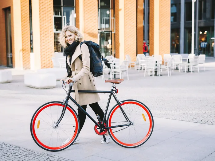 RS-blog-Fiets-van-de-zaak-en-btw Een vrouw met blonde, warrige krullen en een stralende glimlach loopt met haar stijlvolle fixed-gear-fiets met rode velgen door een moderne kantoordistrict.