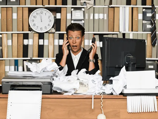 Een verraste zakenvrouw achter een bureau vol met gekreukelde papieren.