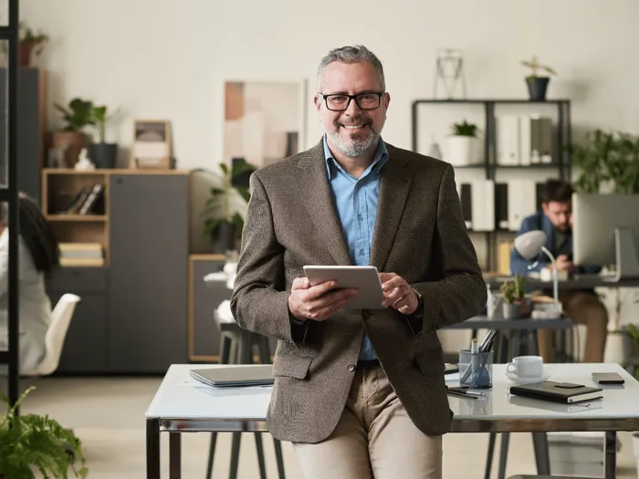 RS-blog-estate-planning Een man in een bruine blazer staat in een licht kantoor met een tablet in zijn hand, leunt tegen een bureau en glimlacht naar de camera.