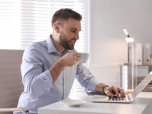 Een persoon in een blauw overhemd zit achter een laptop te werken en drinkt uit een witte mok aan een bureau, met op de achtergrond een moderne lamp en jaloezieën.